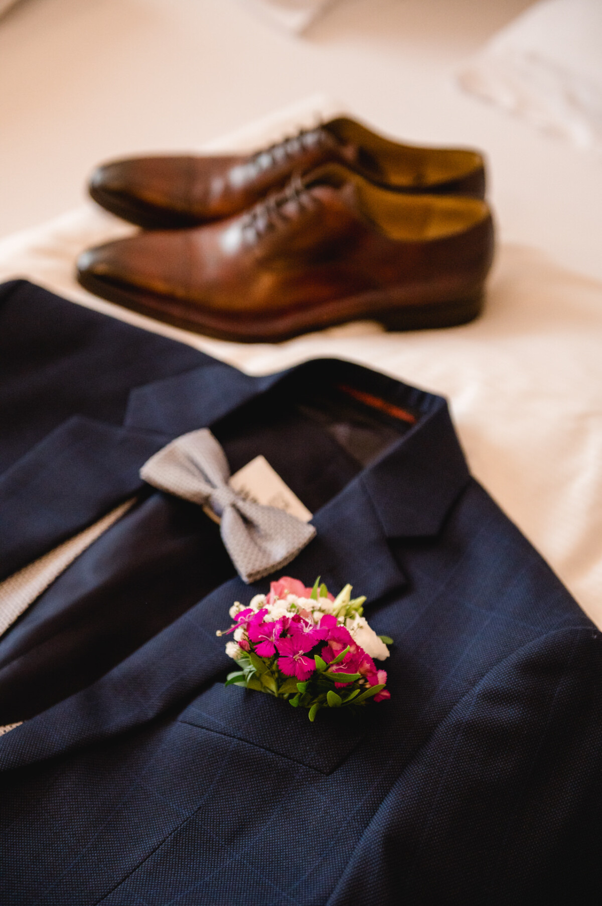 christian-lorenz.net Ein marineblauer Anzug mit grauer Fliege liegt neben braunen Lederschuhen auf einem Bett. An der Anzugjacke ist eine rosafarbene Ansteckblume befestigt.