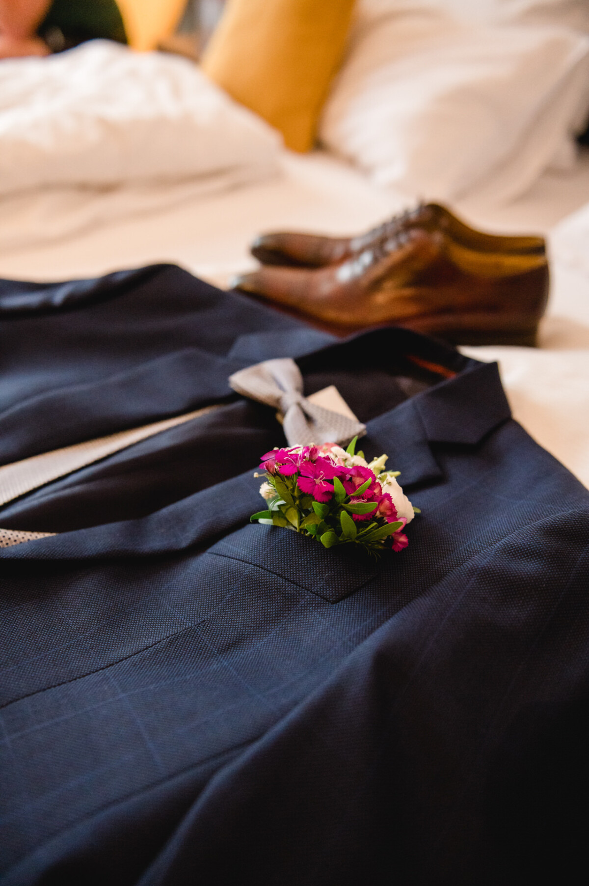 christian-lorenz.net Auf einem Bett liegt ein marineblauer Anzug mit grauer Krawatte und rosa Ansteckblume. Im Hintergrund sind braune Abendschuhe zu sehen.