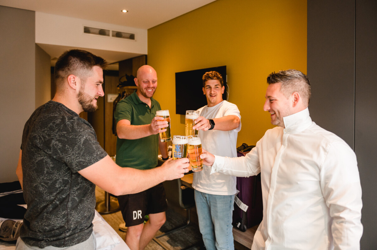 christian-lorenz.net Vier Männer in legerer und halbformeller Kleidung stoßen in einem modernen Raum mit Biergläsern an, in der Ecke stehen Koffer und eine Tasche.