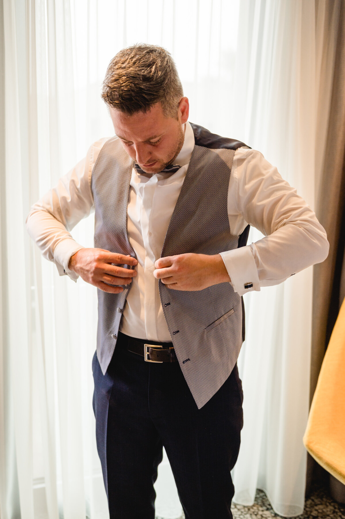 christian-lorenz.net Ein Mann rückt seine graue Weste über einem weißen Hemd und einer Krawatte zurecht und steht vor durchsichtigen Vorhängen.