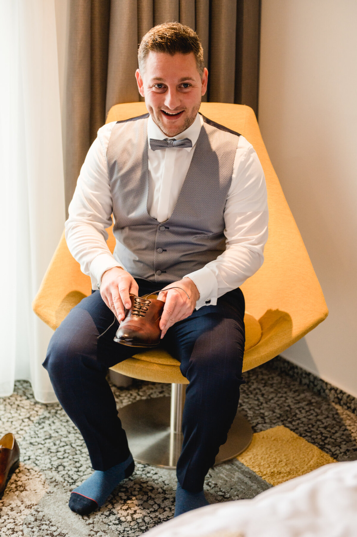 christian-lorenz.net Ein Mann sitzt lächelnd auf einem gelben Stuhl und zieht braune Lederschuhe an. Er trägt eine graue Weste, ein weißes Hemd und eine dunkelblaue Hose. Im Hintergrund sind ein Vorhang und ein Teppichboden zu sehen.