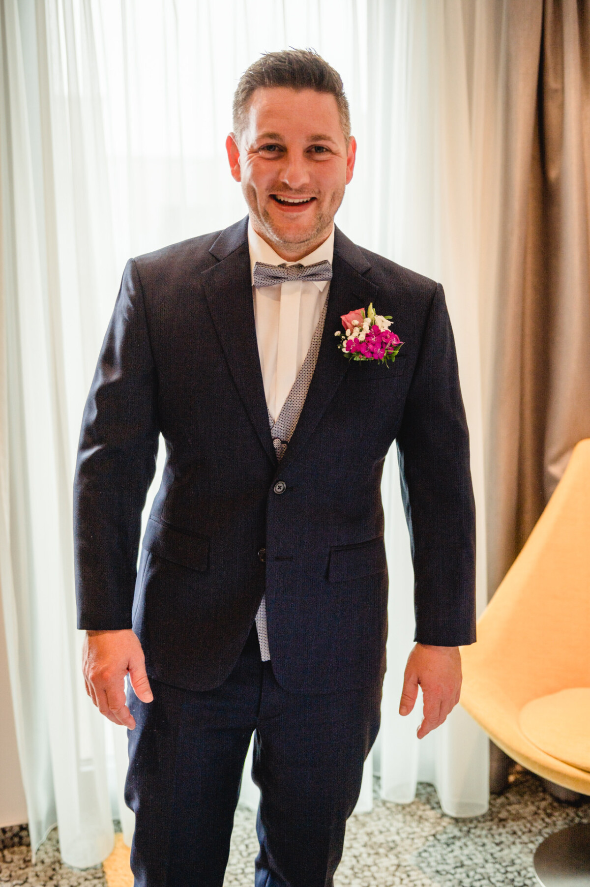 christian-lorenz.net Ein Mann in einem dunklen Anzug mit einer lila Ansteckblume steht lächelnd in einem Raum mit einem Fenster und beigen Vorhängen.