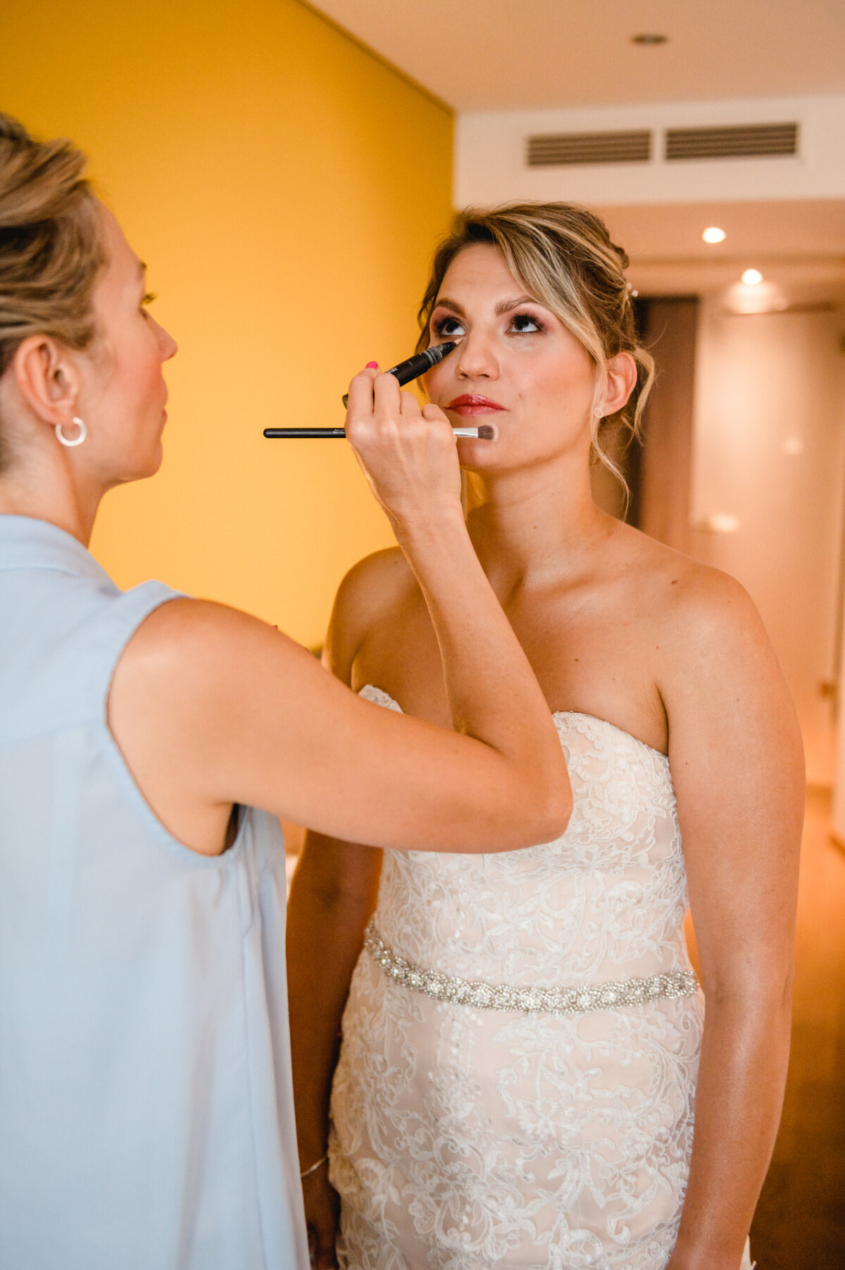 christian-lorenz.net Eine Braut in einem weißen Kleid wird in einem Raum mit einer gelben Wand von einer Frau mit einem Pinsel geschminkt.