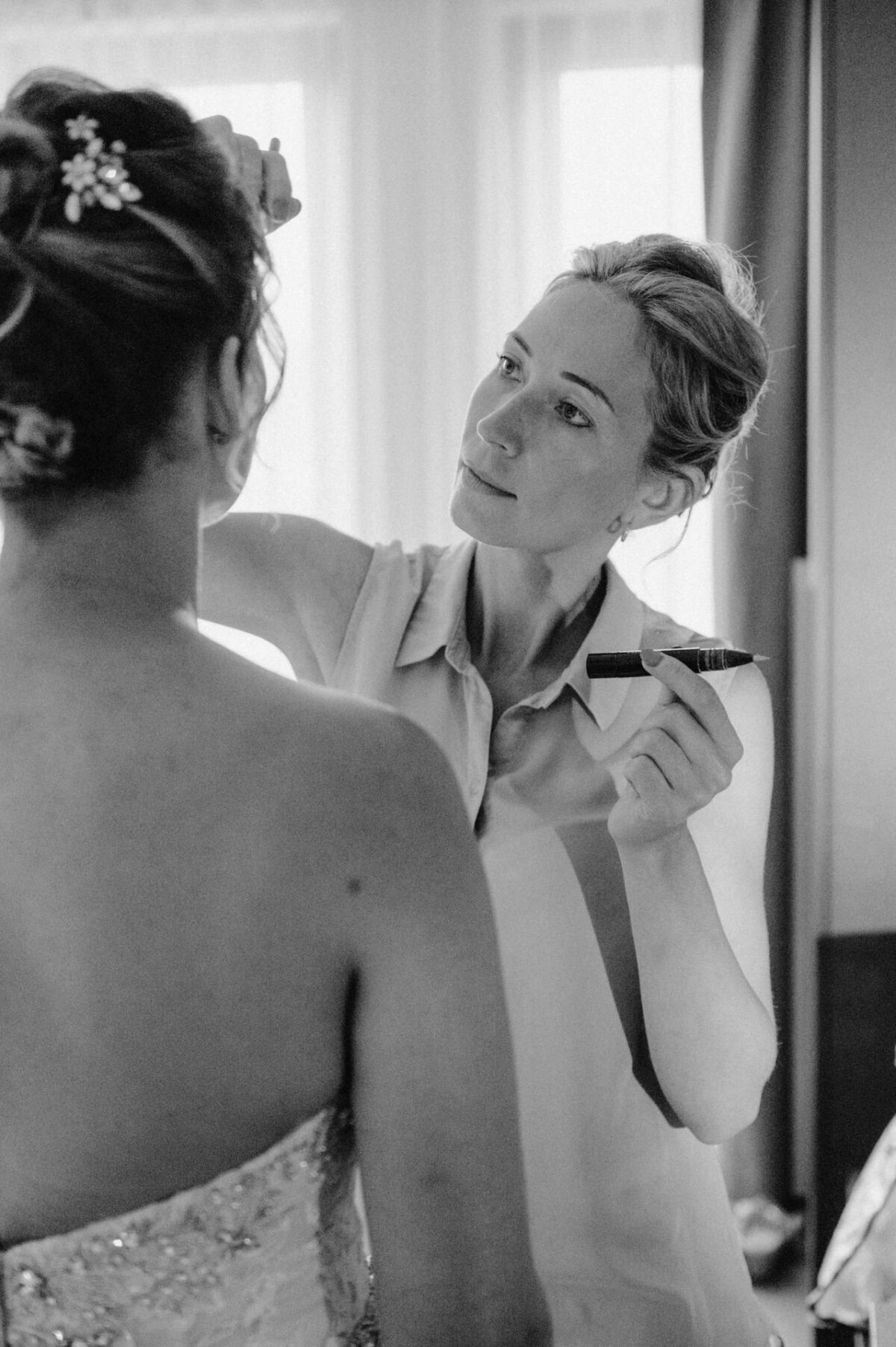 christian-lorenz.net In einem sanft beleuchteten Raum schminkt eine Frau eine andere Frau in einem trägerlosen Kleid mit Perlenverzierung.