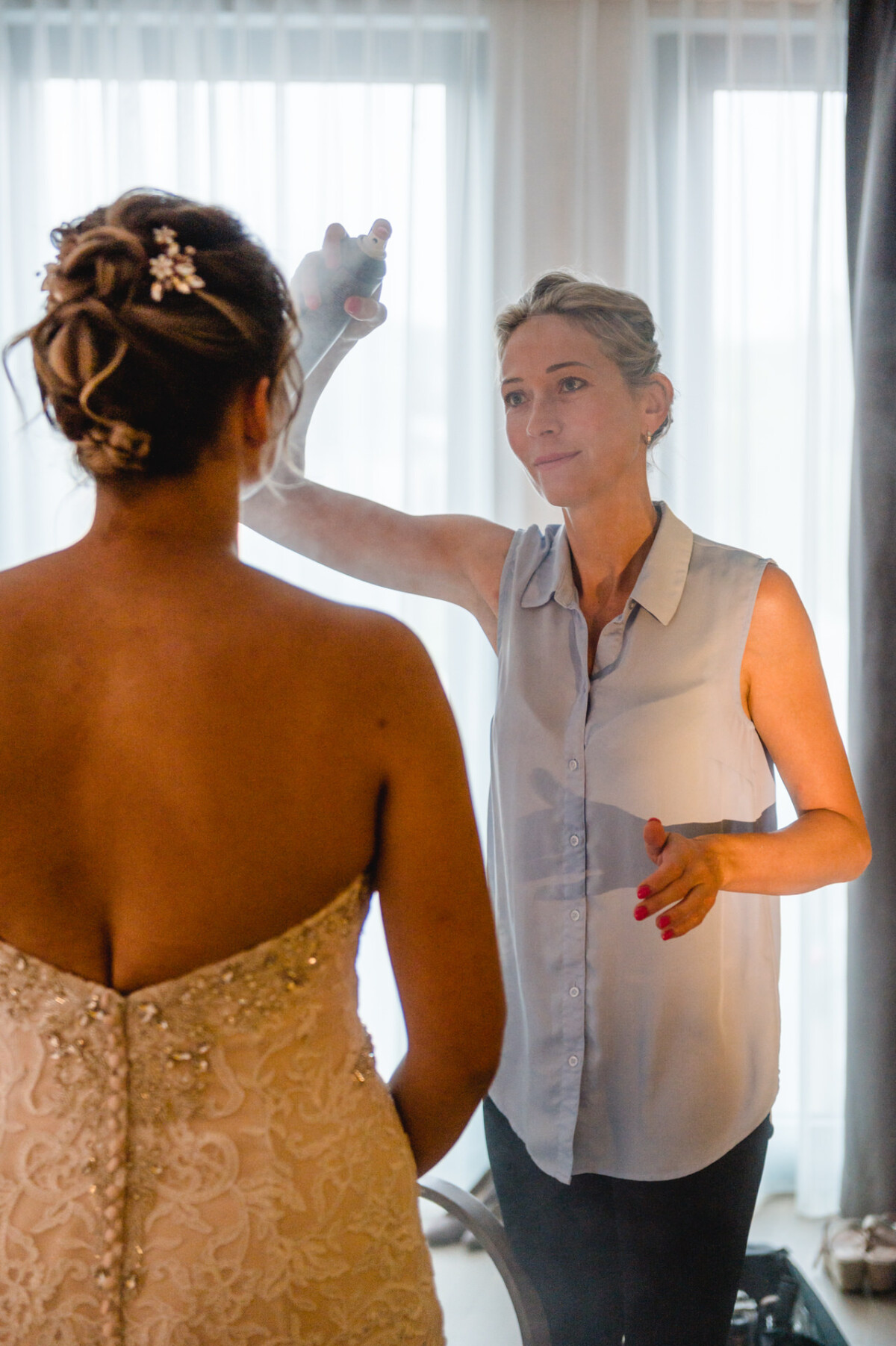 christian-lorenz.net Eine Frau in einer ärmellosen Bluse sprüht Haarspray auf die Rückseite der gestylten Haare einer anderen Frau. Die zweite Frau trägt ein verziertes Brautkleid.