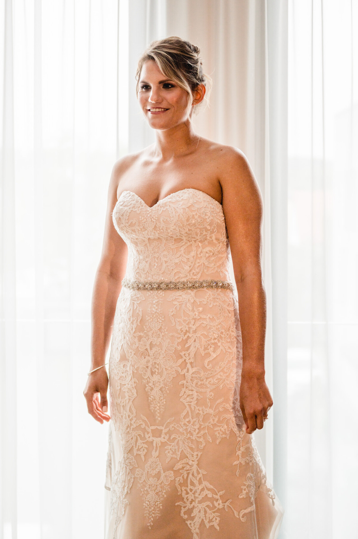 christian-lorenz.net Eine Frau in einem trägerlosen Spitzenbrautkleid steht neben transparenten Vorhängen und lächelt leicht.