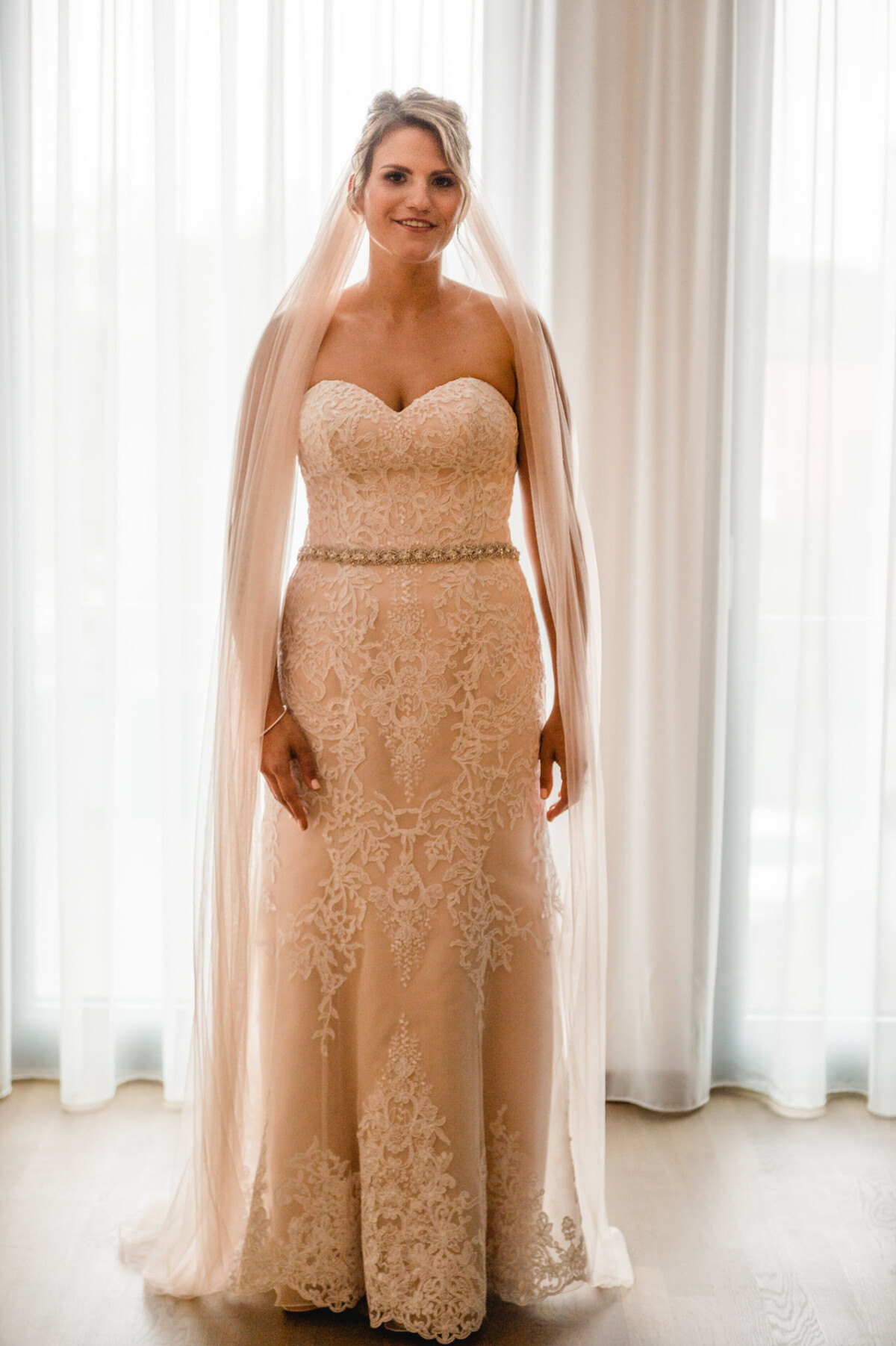 christian-lorenz.net Eine Frau in einem Spitzenbrautkleid steht vor transparenten Vorhängen. Ein langer Schleier liegt über ihren Schultern und sie lächelt in die Kamera.