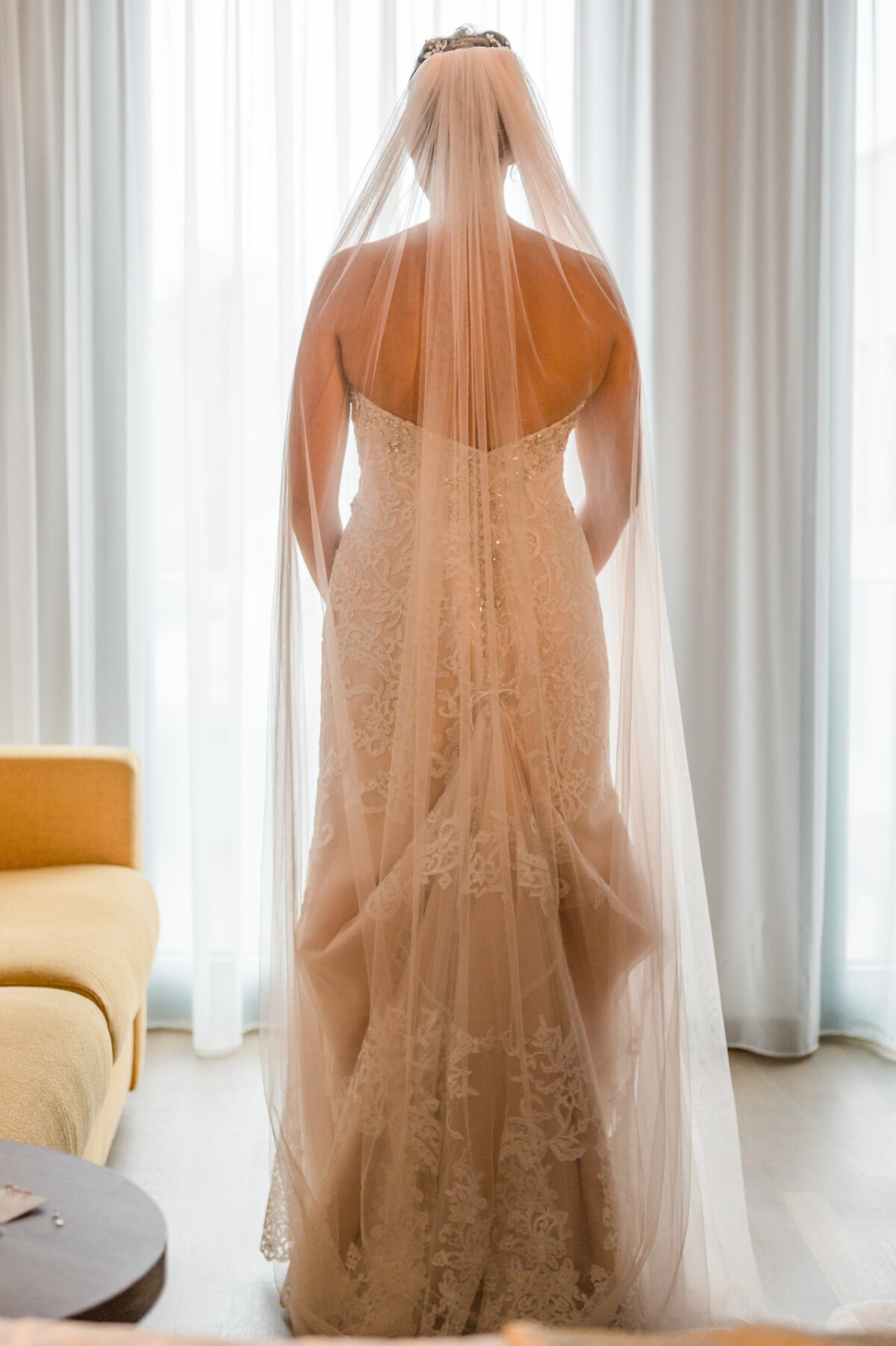 christian-lorenz.net Eine Braut in einem weißen Spitzenbrautkleid und Schleier steht vor einem Fenster. Sie sitzt neben einem gelben Sofa, durch transparente Vorhänge fällt Licht.