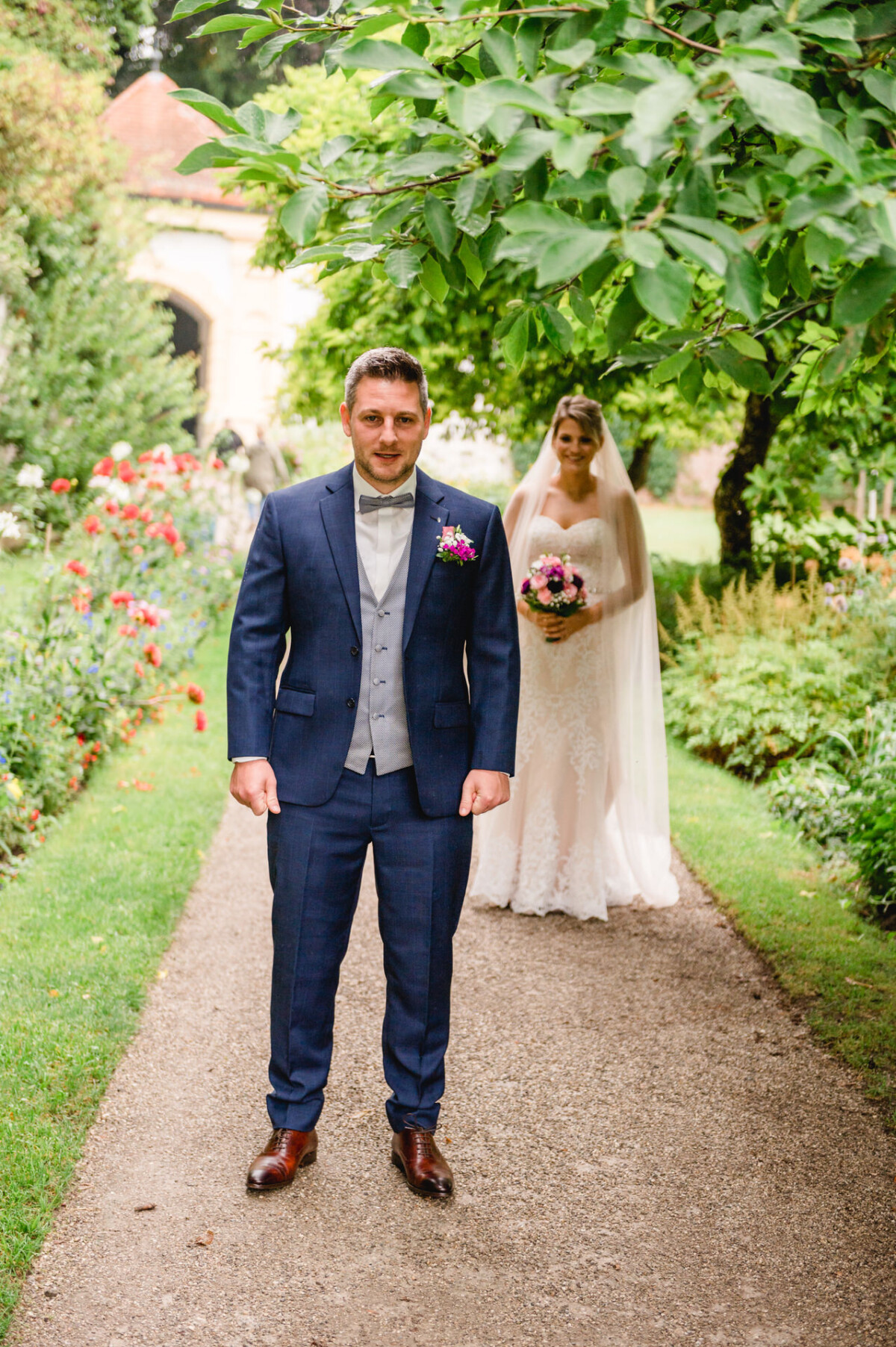 christian-lorenz.net Ein Bräutigam im blauen Anzug steht auf einem Gartenweg, hinter ihm hält eine Braut im weißen Kleid und Schleier einen Blumenstrauß.
