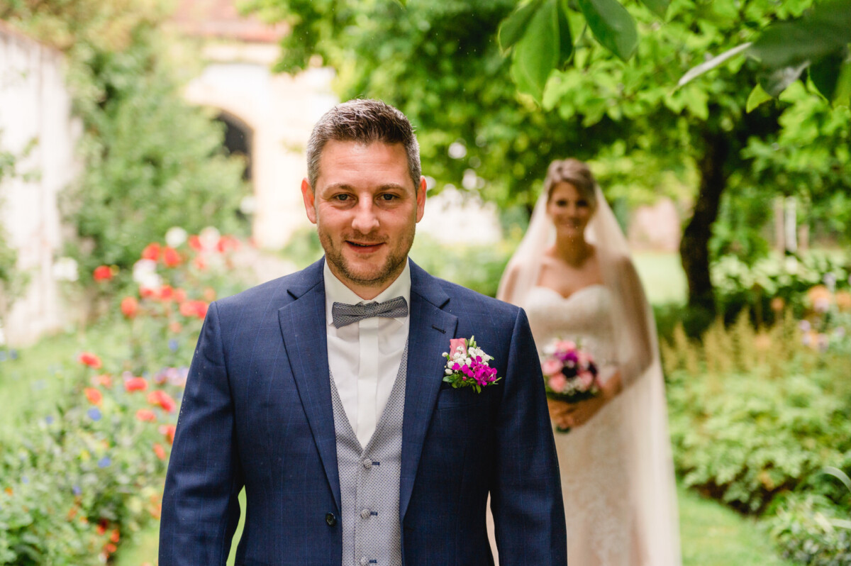 christian-lorenz.net Ein Bräutigam im blauen Anzug steht in einem Garten, im Hintergrund ist eine Braut zu sehen, die einen Blumenstrauß hält und ein weißes Kleid und einen Schleier trägt.