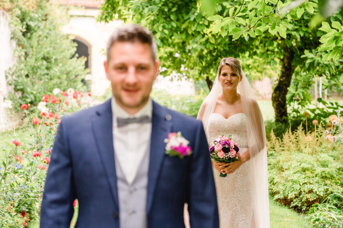christian-lorenz.net Im Vordergrund steht ein Bräutigam im blauen Anzug, während im Hintergrund eine Braut im weißen Kleid und Schleier einen rosa Blumenstrauß hält, umgeben von Grünpflanzen und Blumen.
