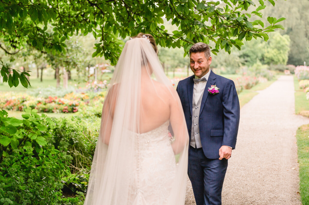 christian-lorenz.net Ein Bräutigam im blauen Anzug reagiert mit einem Lächeln, als er seine Braut im Brautkleid zum ersten Mal sieht. Sie befinden sich im Freien in einer Gartenanlage mit einem Weg und viel Grün drumherum.
