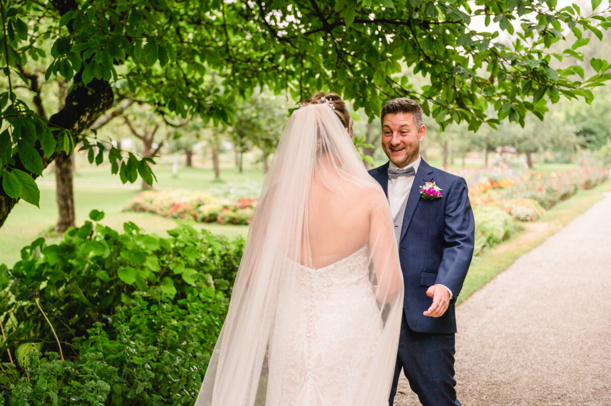 christian-lorenz.net Eine Braut in einem weißen Kleid und Schleier nähert sich einem lächelnden Mann im Anzug mit einer Ansteckblume, der auf einem Gartenweg mit Bäumen und Blumen im Hintergrund steht.