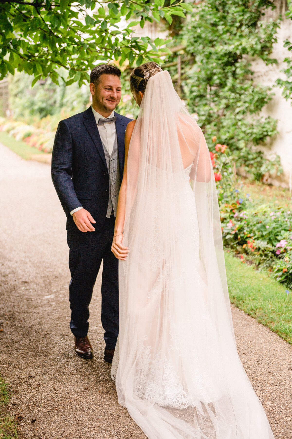 christian-lorenz.net Ein Bräutigam im dunklen Anzug lächelt eine Braut im weißen Kleid mit langem Schleier an. Sie stehen auf einem Kiesweg, umgeben von Grün und bunten Blumen.