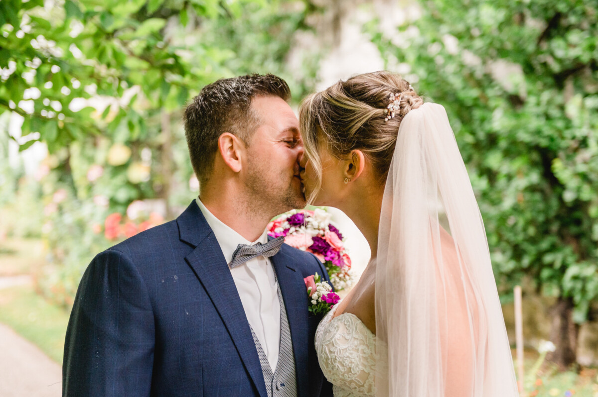 christian-lorenz.net Braut und Bräutigam küssen sich im Freien. Die Braut trägt ein weißes Kleid und einen Schleier und hält einen Blumenstrauß in der Hand. Der Bräutigam trägt einen blauen Anzug. Im Hintergrund sind Grünpflanzen und Blumen zu sehen.
