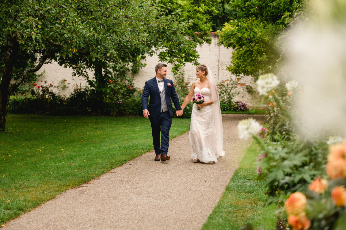 christian-lorenz.net Eine Braut und ein Bräutigam gehen Hand in Hand einen Gartenweg entlang, umgeben von Grün und Blumen.