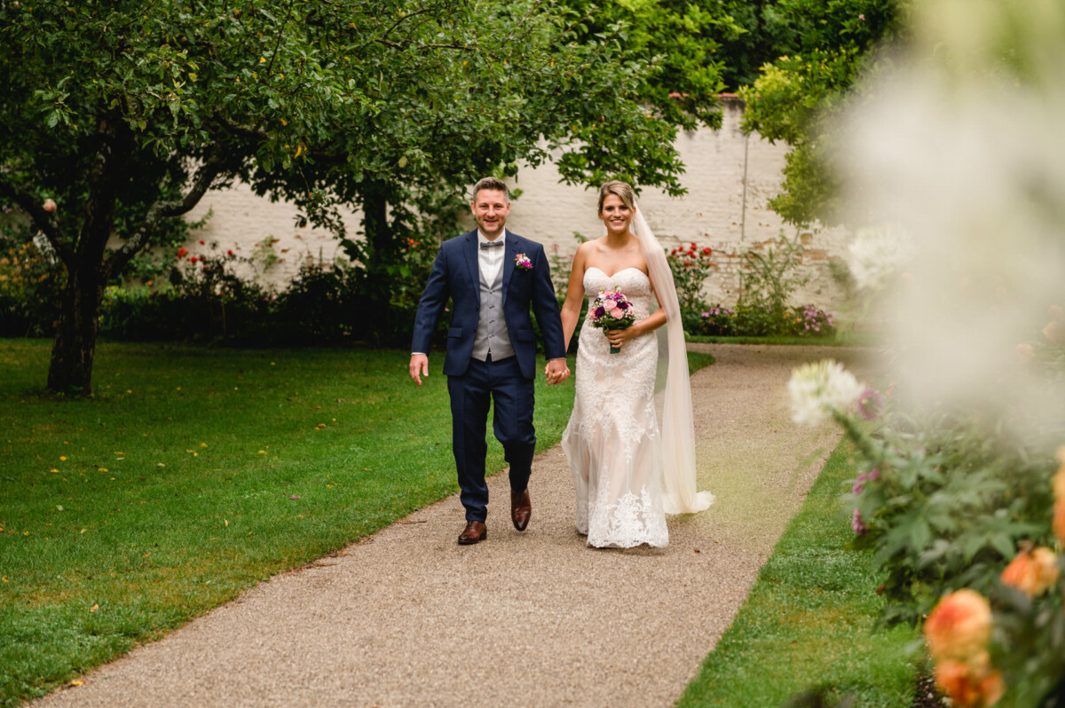 christian-lorenz.net Braut und Bräutigam lächeln, während sie Hand in Hand einen Gartenweg entlanggehen. Die Braut hält einen Blumenstrauß in der Hand, und sie sind von viel Grün umgeben.