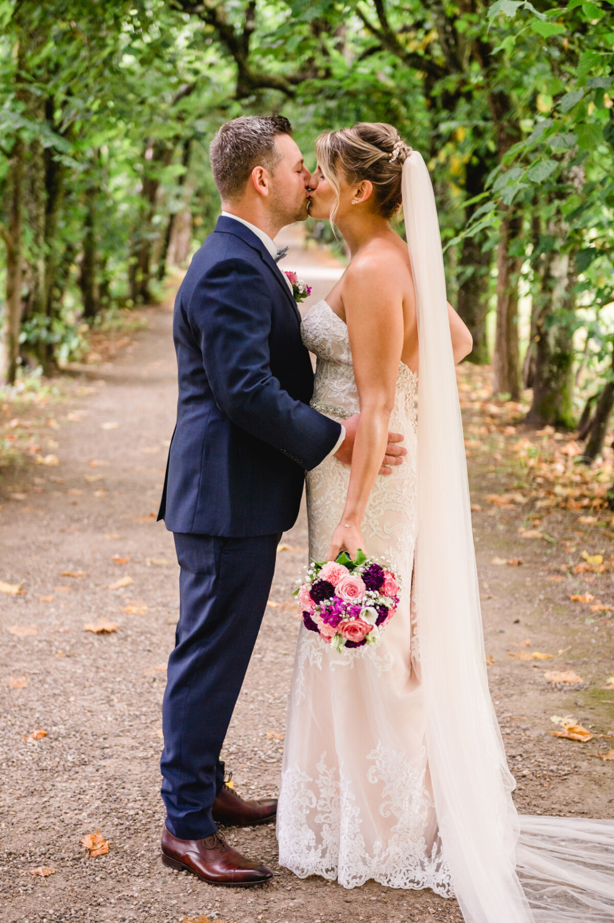 christian-lorenz.net Braut und Bräutigam in festlicher Kleidung küssen sich auf einem von Bäumen gesäumten Weg. Die Braut hält einen Blumenstrauß und trägt einen langen Schleier.
