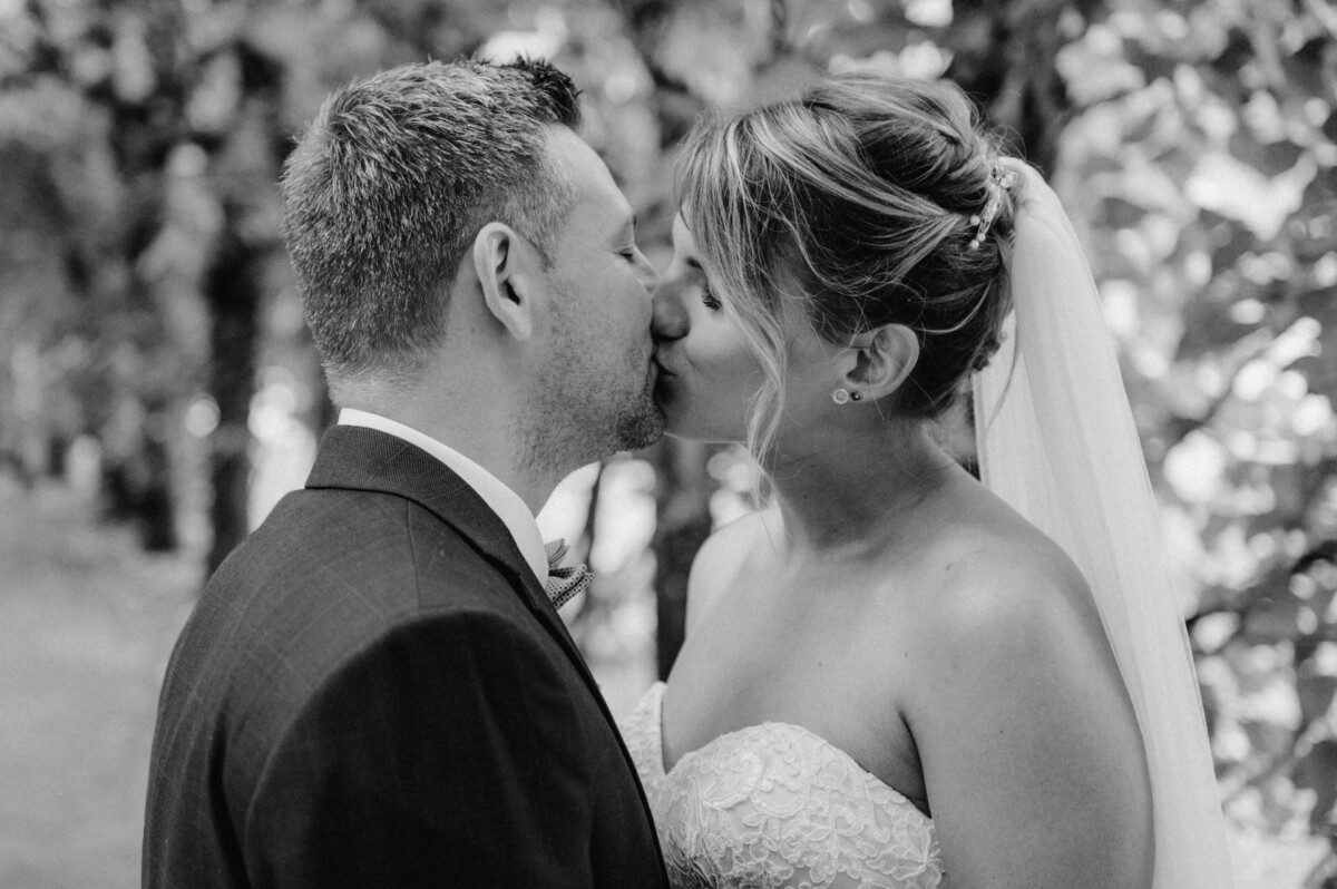 christian-lorenz.net Braut und Bräutigam küssen sich im Freien. Die Braut trägt ein trägerloses Kleid und einen Schleier, der Bräutigam einen dunklen Anzug. Im Hintergrund sind Bäume zu sehen.