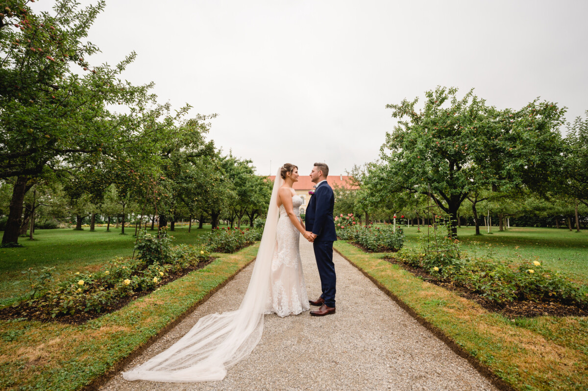 christian-lorenz.net An einem bewölkten Tag stehen Braut und Bräutigam einander gegenüber und halten sich an den Händen auf einem von Bäumen und Grün gesäumten Kiesweg.