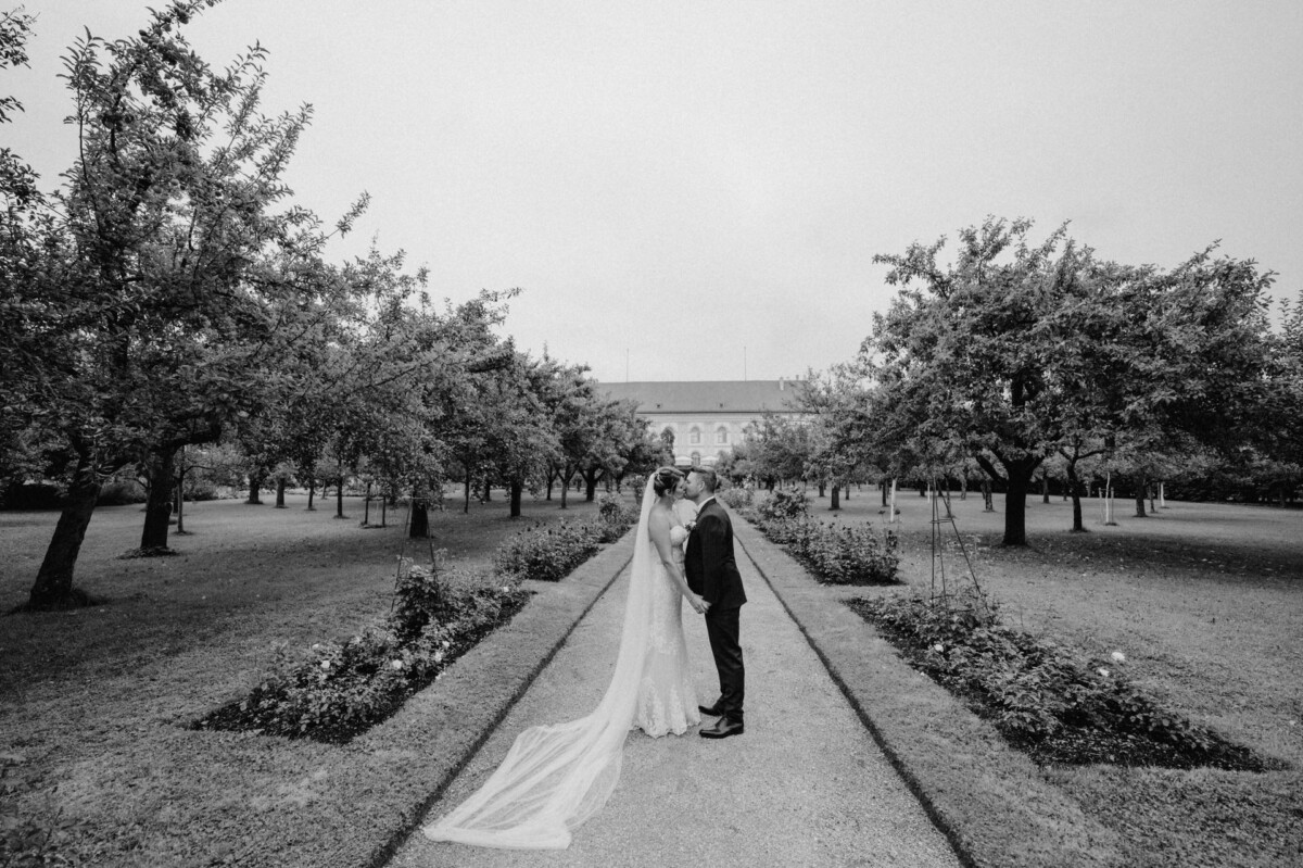 christian-lorenz.net Braut und Bräutigam küssen sich auf einem Weg zwischen Bäumen in einer Gartenlandschaft. Der lange Schleier der Braut weht hinter ihnen auf dem Kiesweg her. Die Szene ist in Schwarzweiß gehalten.