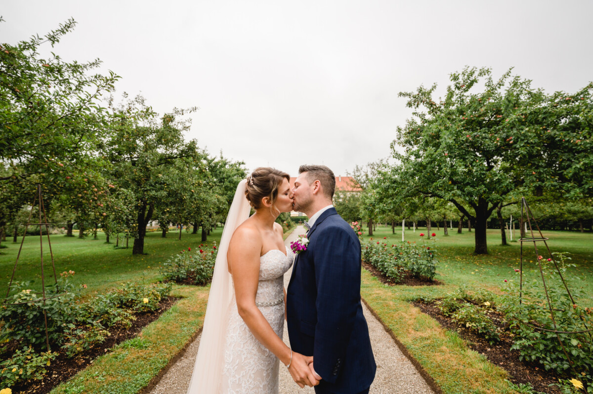 christian-lorenz.net Braut und Bräutigam küssen sich an einem bewölkten Tag auf einem von Bäumen und blühenden Pflanzen gesäumten Gartenweg.