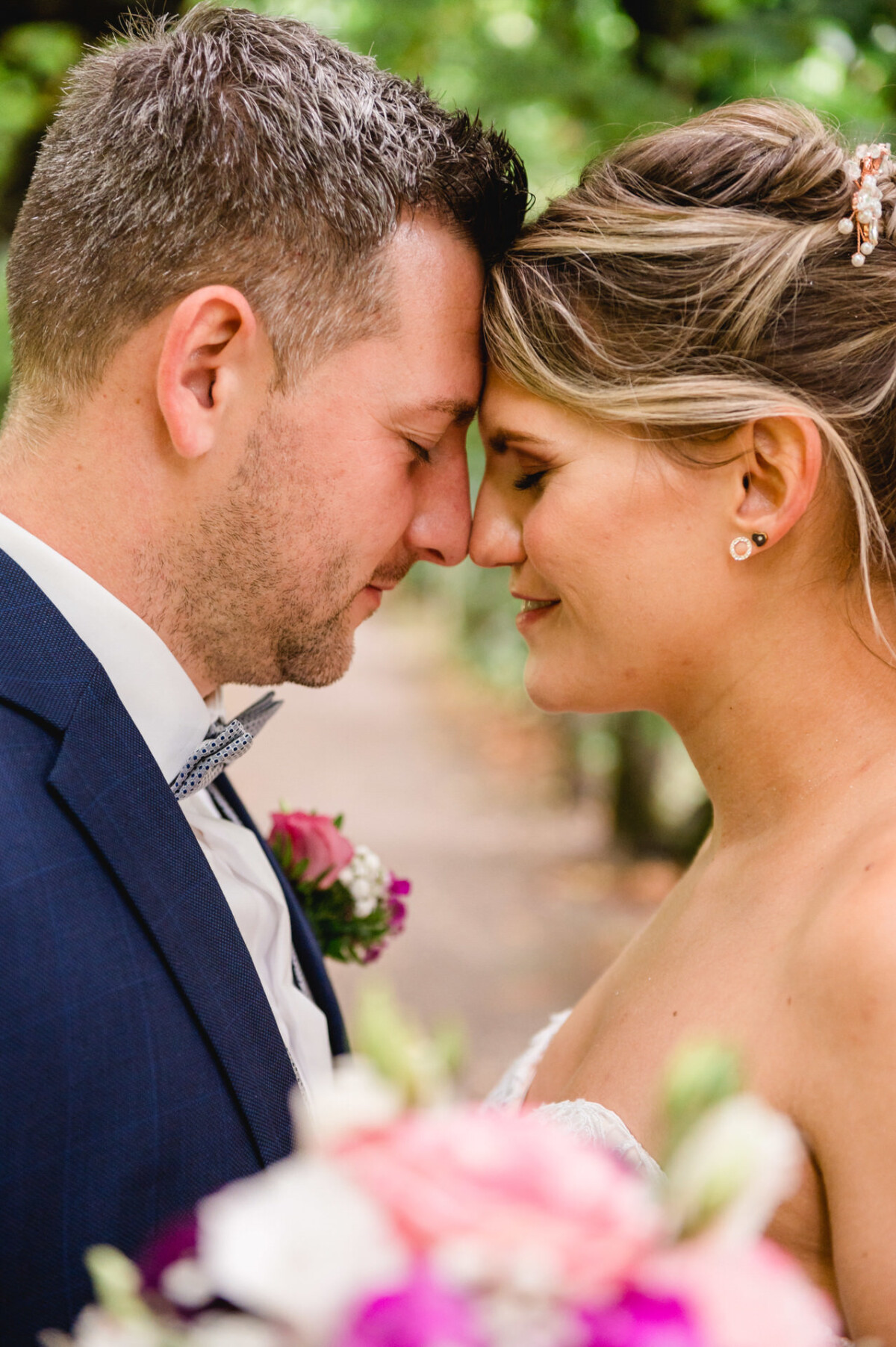 christian-lorenz.net Eine Braut und ein Bräutigam berühren sanft ihre Stirn, haben die Augen geschlossen, sind von Grün umgeben und halten einen Strauß rosa und weißer Blumen in den Händen.