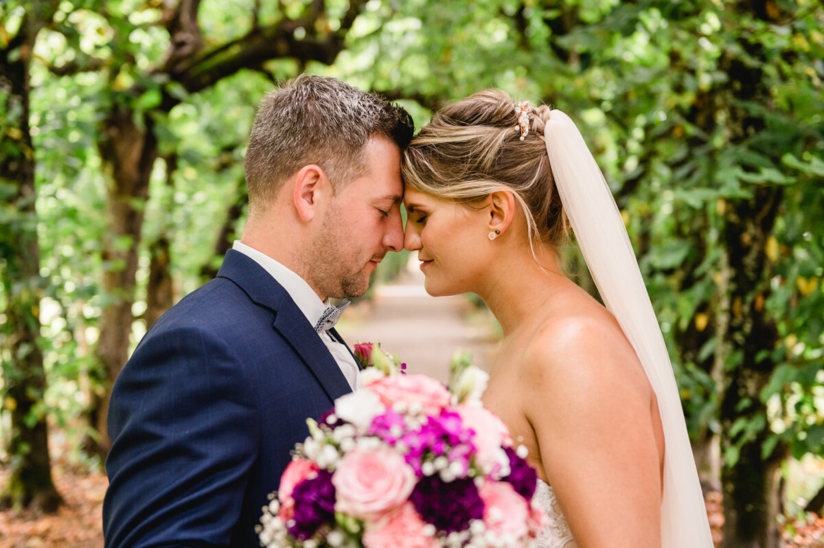 christian-lorenz.net Braut und Bräutigam stehen eng beieinander in einer üppigen, grünen Umgebung und berühren sanft ihre Stirn. Die Braut hält einen Strauß rosa und lila Blumen in den Händen.