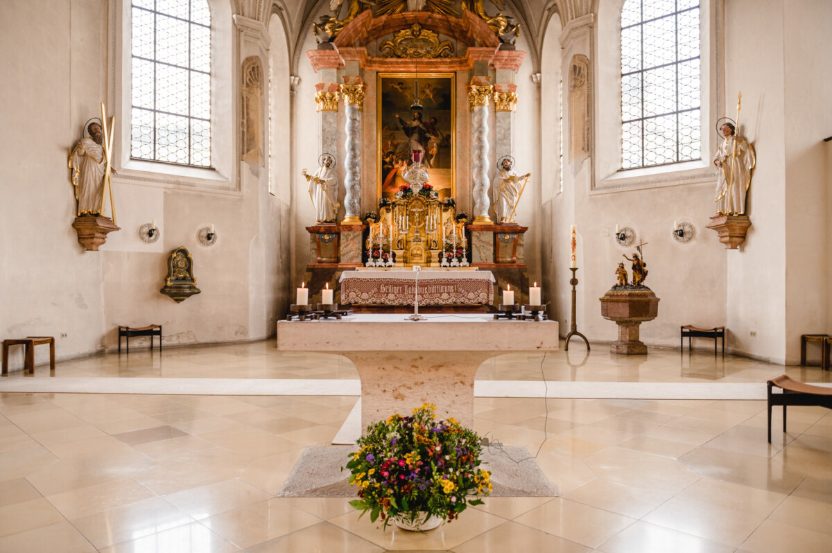 christian-lorenz.net Kirchenaltar mit einem Blumenarrangement im Vordergrund, umgeben von Kerzen und Statuen. Großes Gemälde und kunstvolle Verzierungen im Hintergrund.