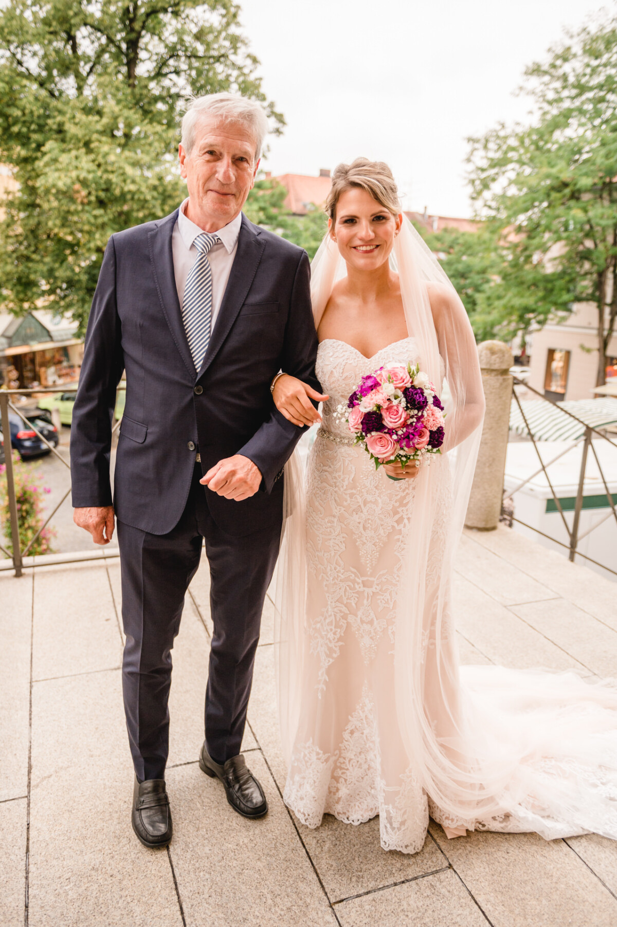 christian-lorenz.net Eine Braut in einem weißen Brautkleid mit einem Blumenstrauß steht Arm in Arm mit einem älteren Mann im Anzug auf einer Steintreppe, im Hintergrund sind Bäume und Gebäude zu sehen.