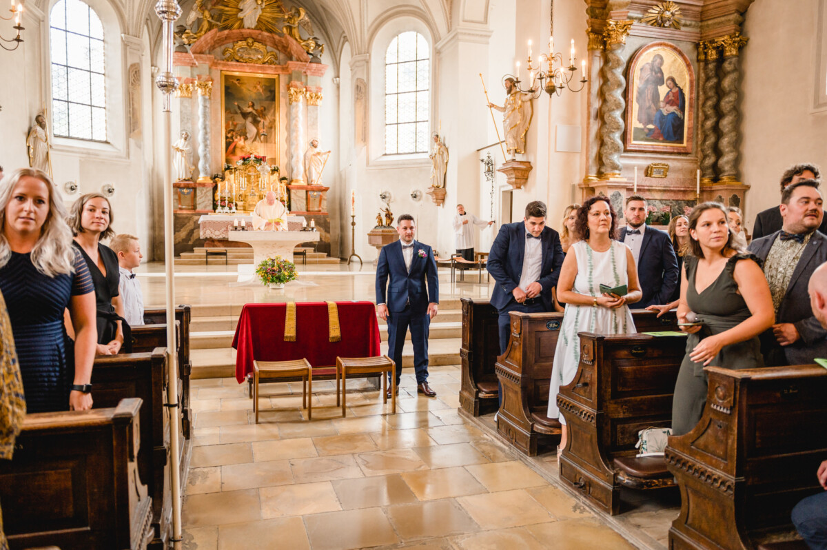 christian-lorenz.net Eine Hochzeitszeremonie in einer Kirche. Die Gäste sitzen in den Kirchenbänken. Der Bräutigam steht am Altar neben einem rot gedeckten Tisch mit Stühlen. Der Altar ist mit religiösen Kunstwerken und Dekorationen geschmückt.