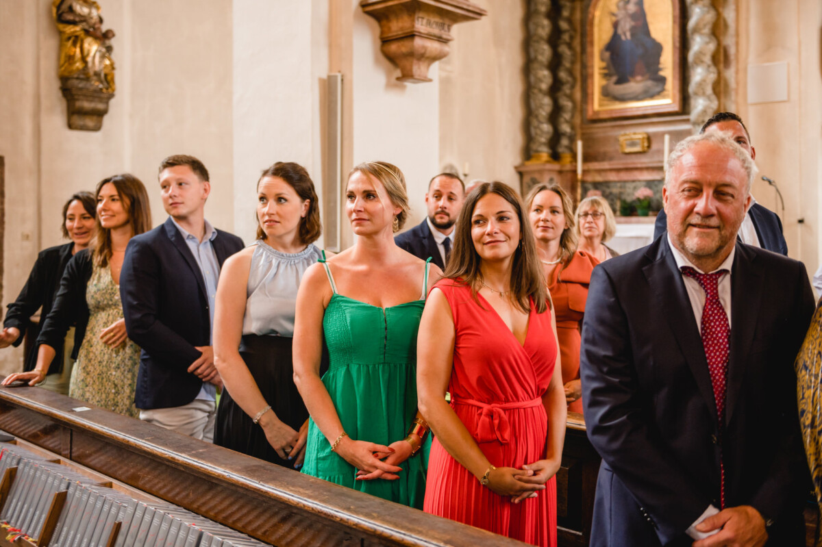 christian-lorenz.net Eine Gruppe von Menschen in Abendgarderobe steht in den Kirchenbänken einer Kirche, wobei einige nach vorne blicken und andere zur Seite blicken.