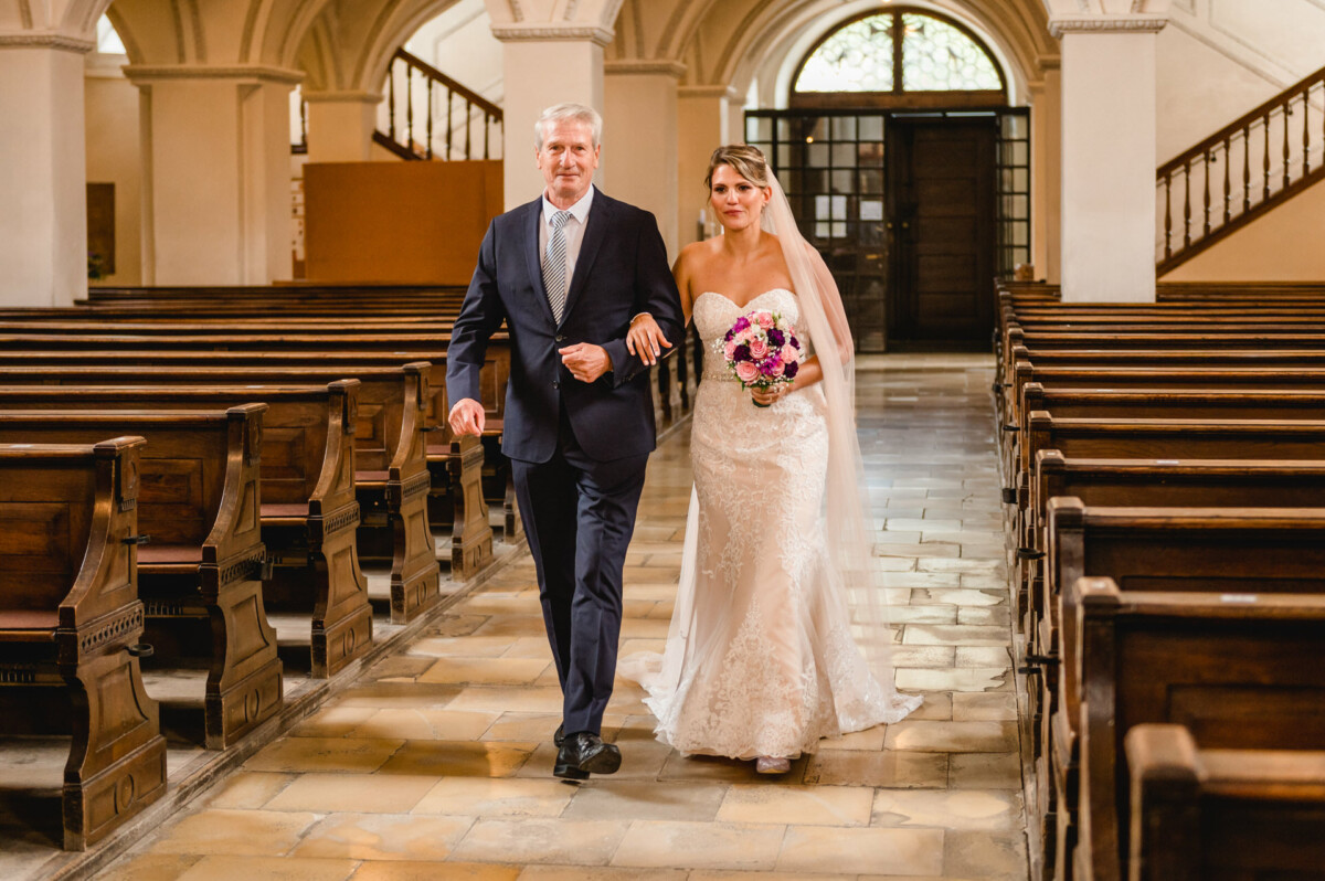 christian-lorenz.net Eine Braut in einem weißen Kleid und einem Blumenstrauß in der Hand geht Arm in Arm mit einem älteren Mann im Anzug den Kirchengang entlang.