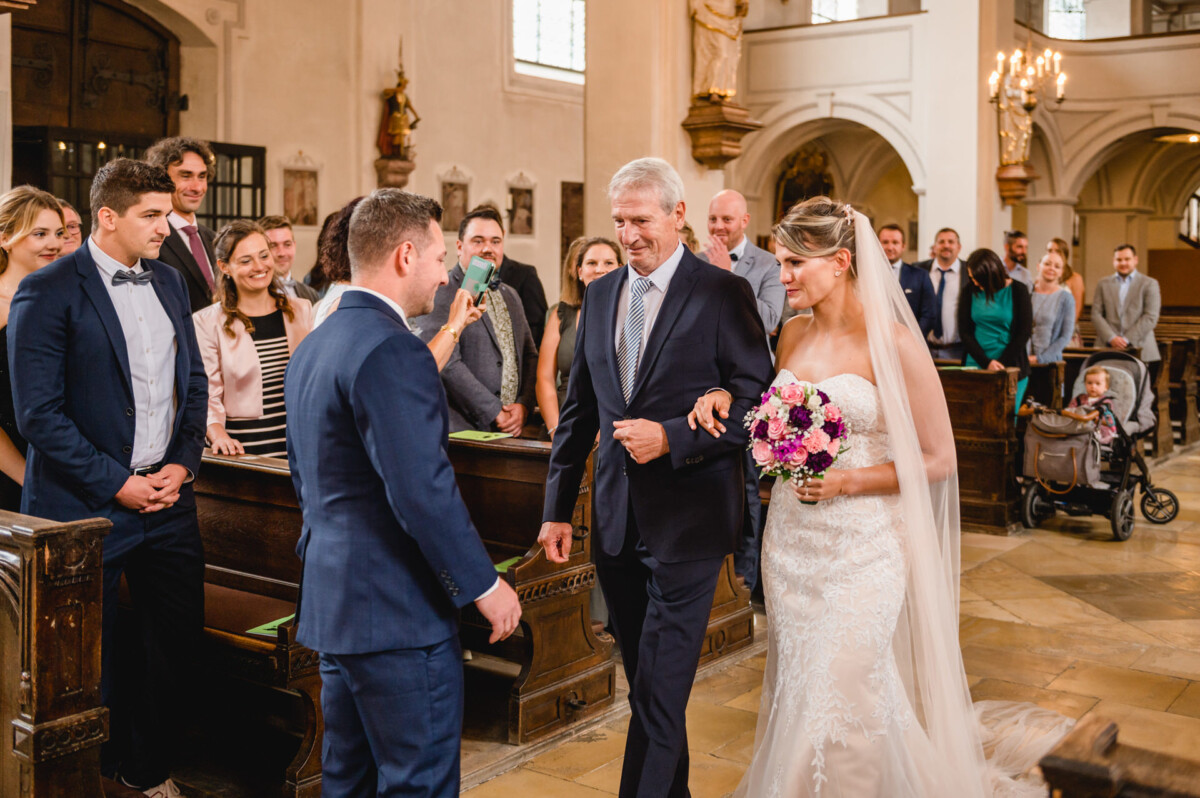 christian-lorenz.net Die Braut im weißen Kleid schreitet mit einem älteren Mann und einem Blumenstrauß in der Hand zum Altar. Der Bräutigam im blauen Anzug steht daneben. Die Gäste schauen in einer kirchlichen Umgebung mit Holzbänken und Kerzen zu.