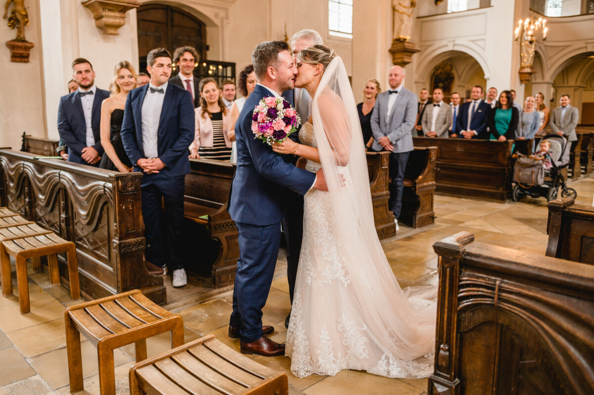 christian-lorenz.net Eine Braut und ein Bräutigam küssen sich in einer Kirche, umgeben von Hochzeitsgästen, die in den Kirchenbänken sitzen und stehen, und im Blickfeld liegt ein Blumenstrauß.