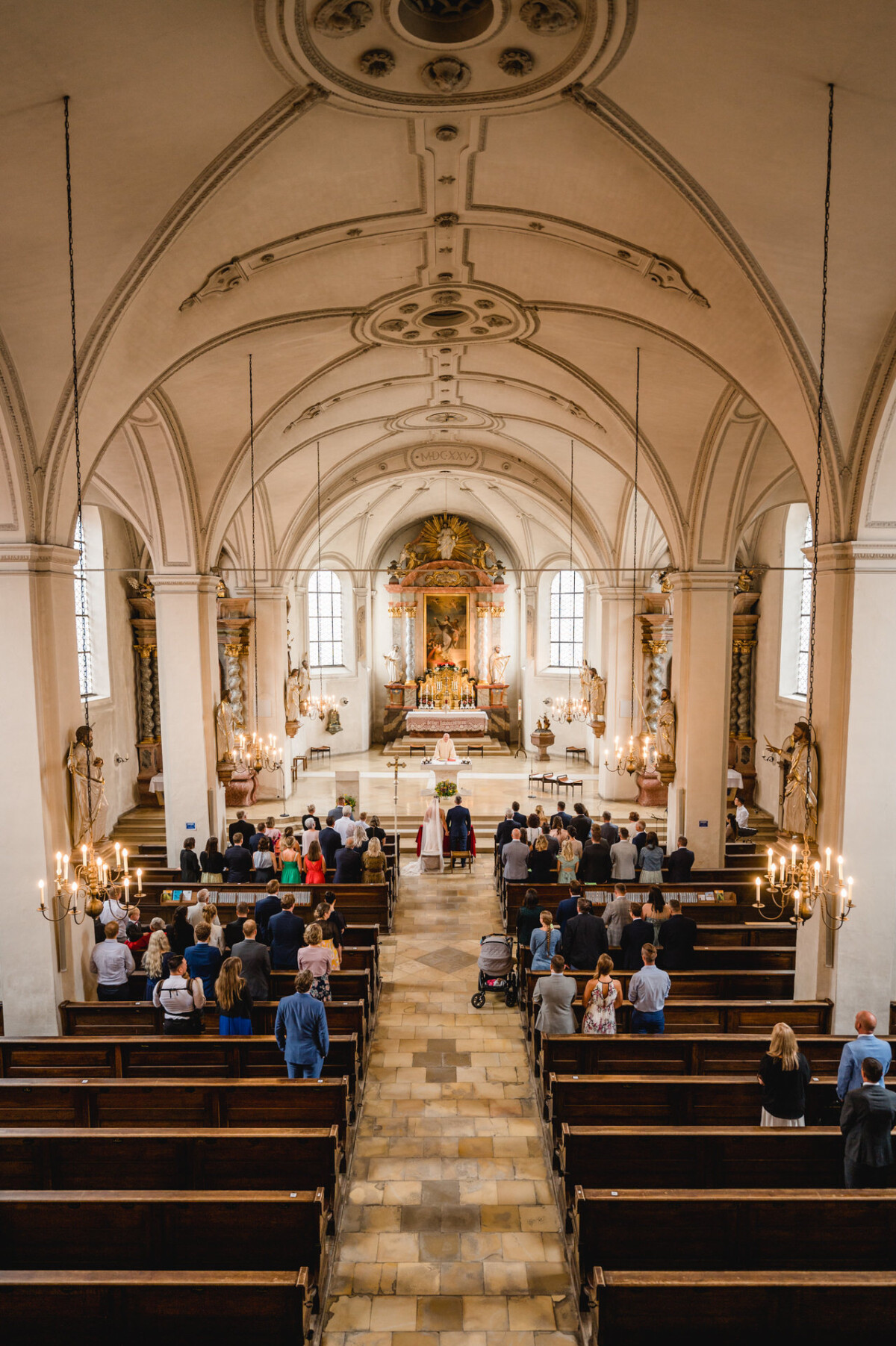 christian-lorenz.net Menschen versammeln sich in einer Kirche, sitzen und stehen in Reihen von Holzbänken vor einem reich verzierten Altar, von dessen Decke Kronleuchter hängen.