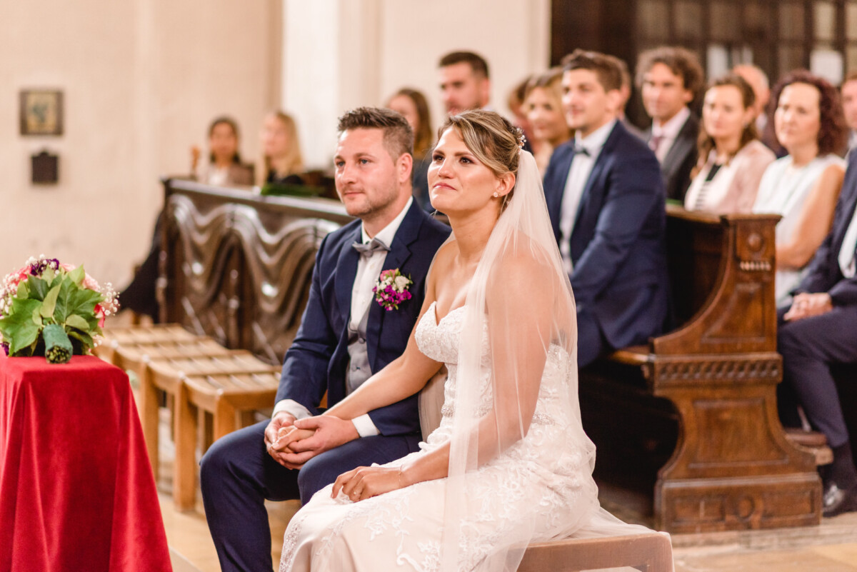 christian-lorenz.net Eine Braut und ein Bräutigam sitzen bei einer Hochzeitszeremonie in einer Kirche Händchen haltend, umgeben von Gästen.