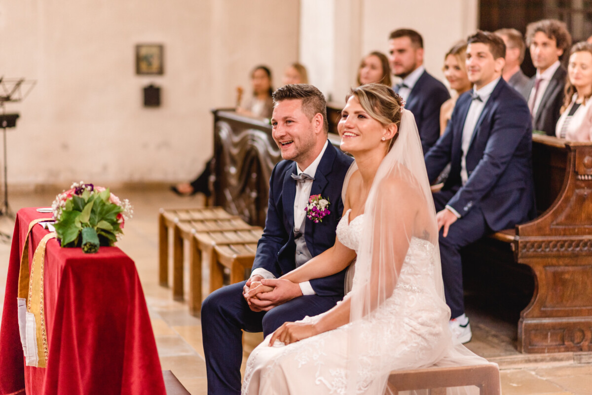 christian-lorenz.net Braut und Bräutigam sitzen während einer Hochzeitszeremonie lächelnd am Altar. Im Hintergrund sitzen Gäste.