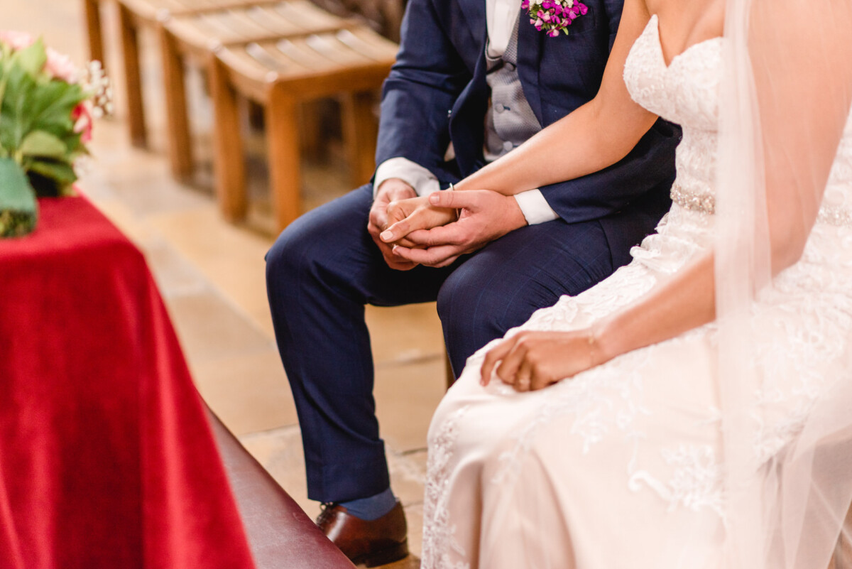 christian-lorenz.net Braut und Bräutigam sitzen während einer Hochzeitszeremonie Händchen haltend da. Der Bräutigam trägt einen dunklen Anzug, während die Braut ein weißes Kleid und einen Schleier trägt.