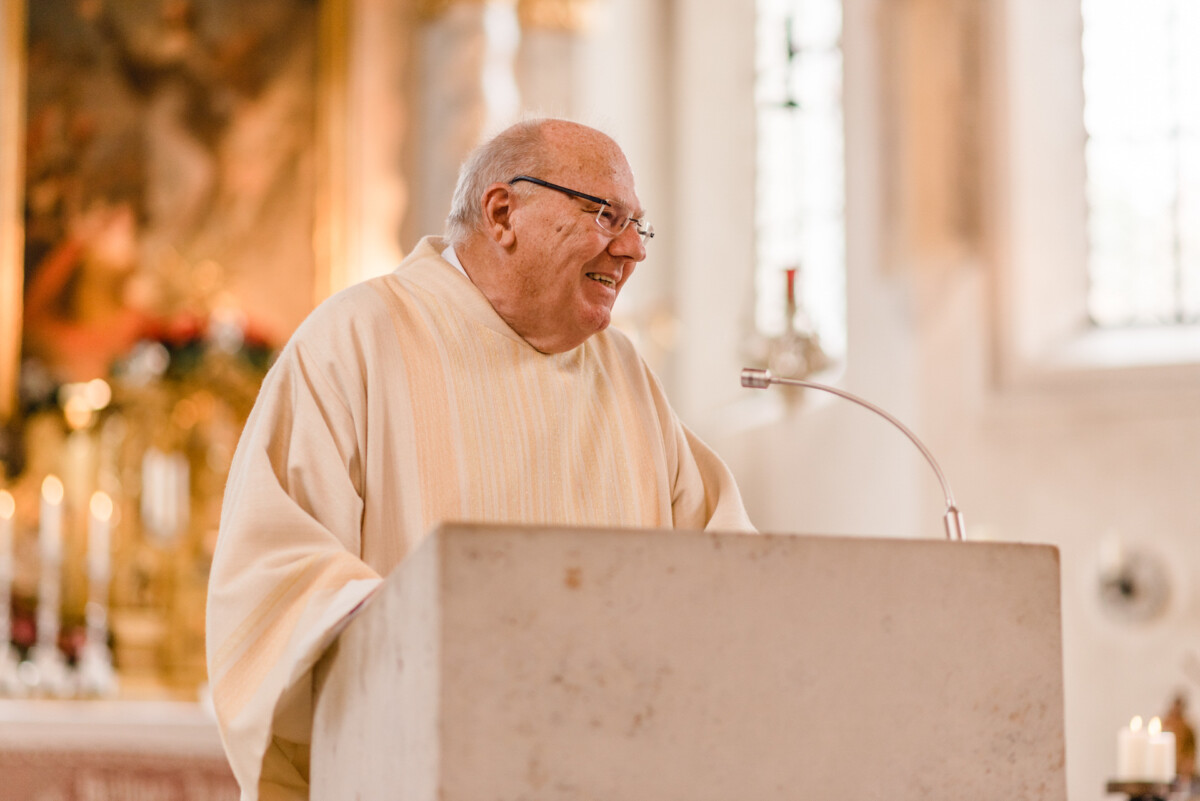 christian-lorenz.net Ein Mann in einem hellen Gewand spricht an einem Podium in einer Kirche mit Kerzen und einem Gemälde im Hintergrund.