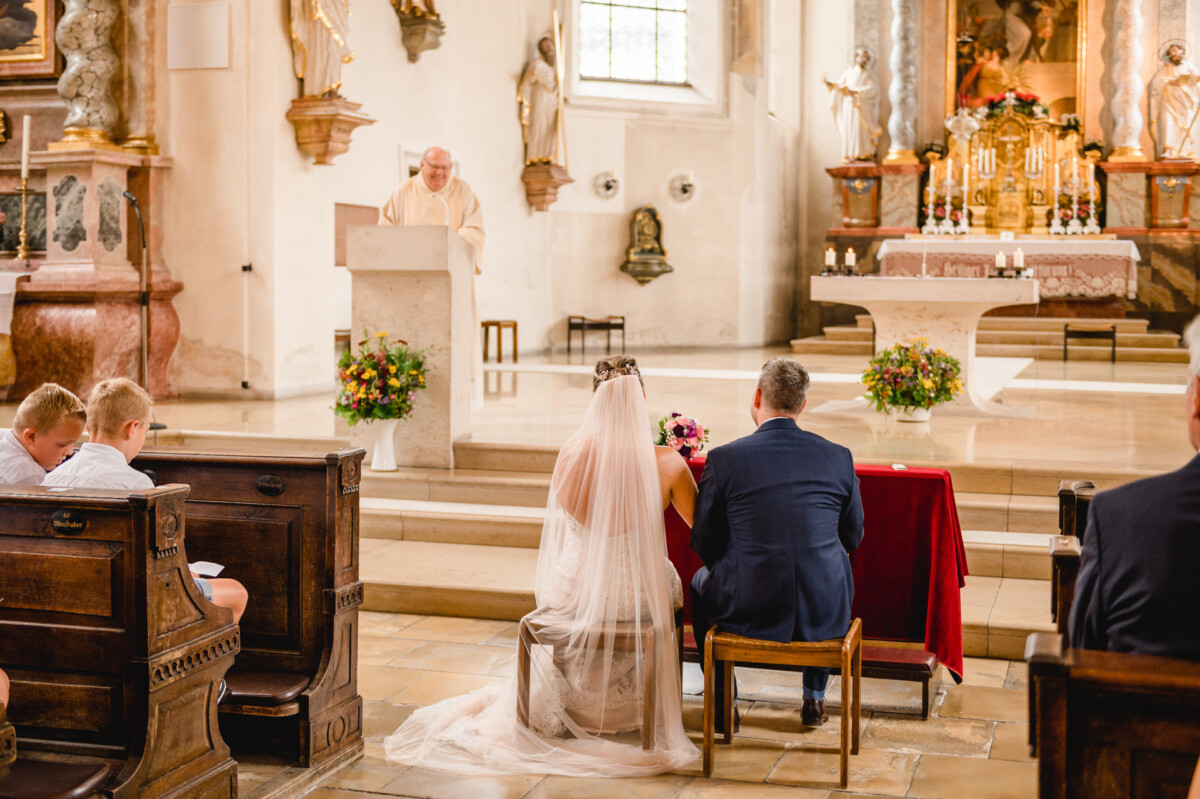 christian-lorenz.net Eine Hochzeitszeremonie in einer Kirche. Braut und Bräutigam sitzen mit Blick zum Altar. Der Geistliche hält die Rede an einem Podium. Die Anwesenden sitzen auf Holzbänken.