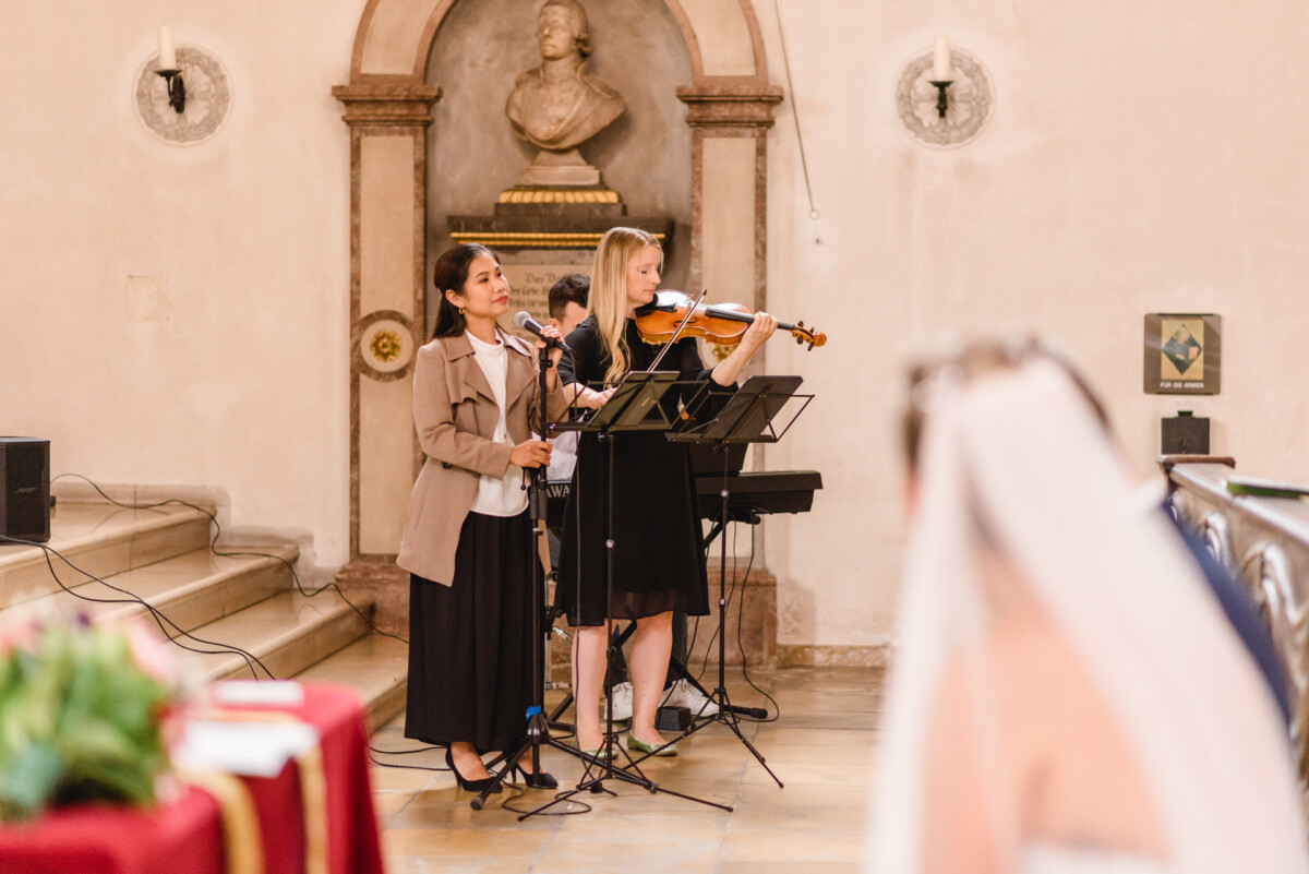 christian-lorenz.net Eine Frau singt in ein Mikrofon, während eine andere in einer Kirche Bratsche spielt. Es findet eine Hochzeitszeremonie statt.