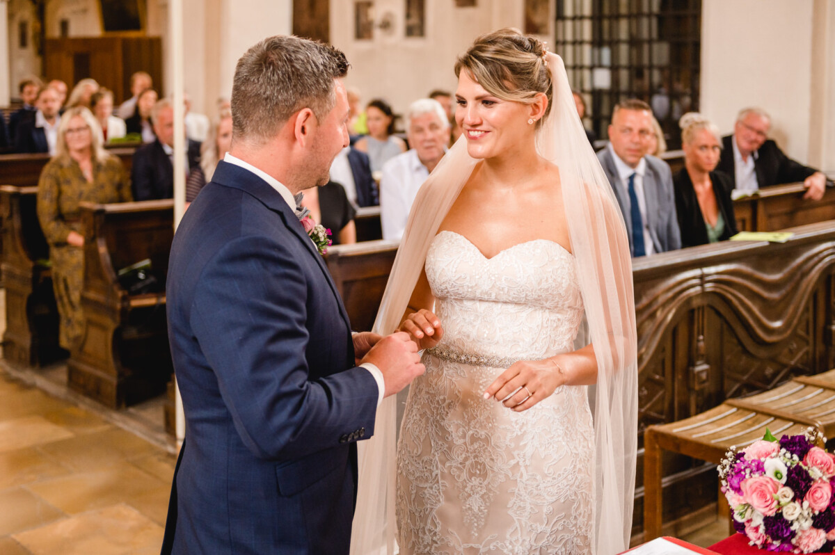 christian-lorenz.net Während der Hochzeitszeremonie stehen Braut und Bräutigam in einer kirchlichen Umgebung am Altar, umgeben von sitzenden Gästen.