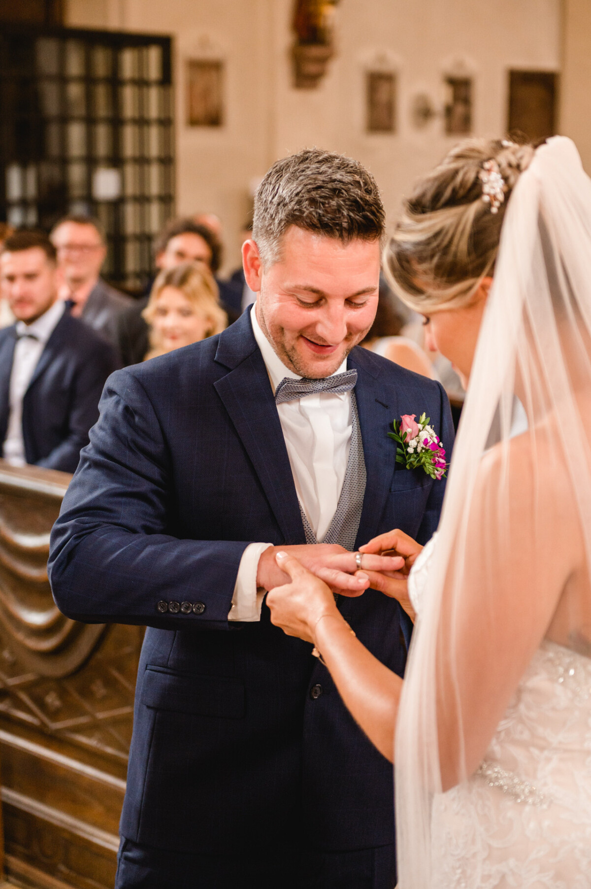 christian-lorenz.net Eine Braut steckt dem Bräutigam während einer Hochzeitszeremonie in einer Kirche einen Ring an den Finger. Im Hintergrund sitzen Gäste in festlicher Kleidung.