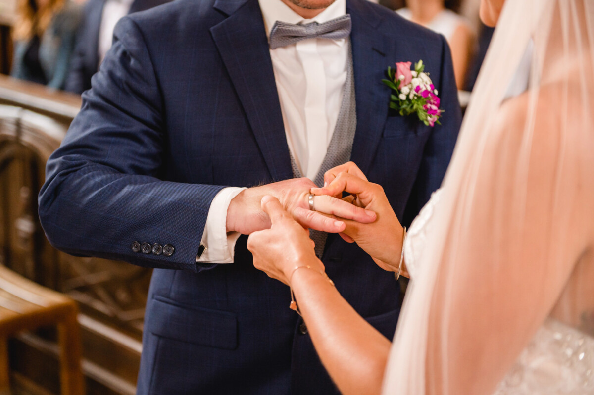 christian-lorenz.net Eine Braut steckt dem Bräutigam während einer Hochzeitszeremonie einen Ring an den Finger. Der Bräutigam trägt einen dunklen Anzug mit Fliege und Ansteckblume.