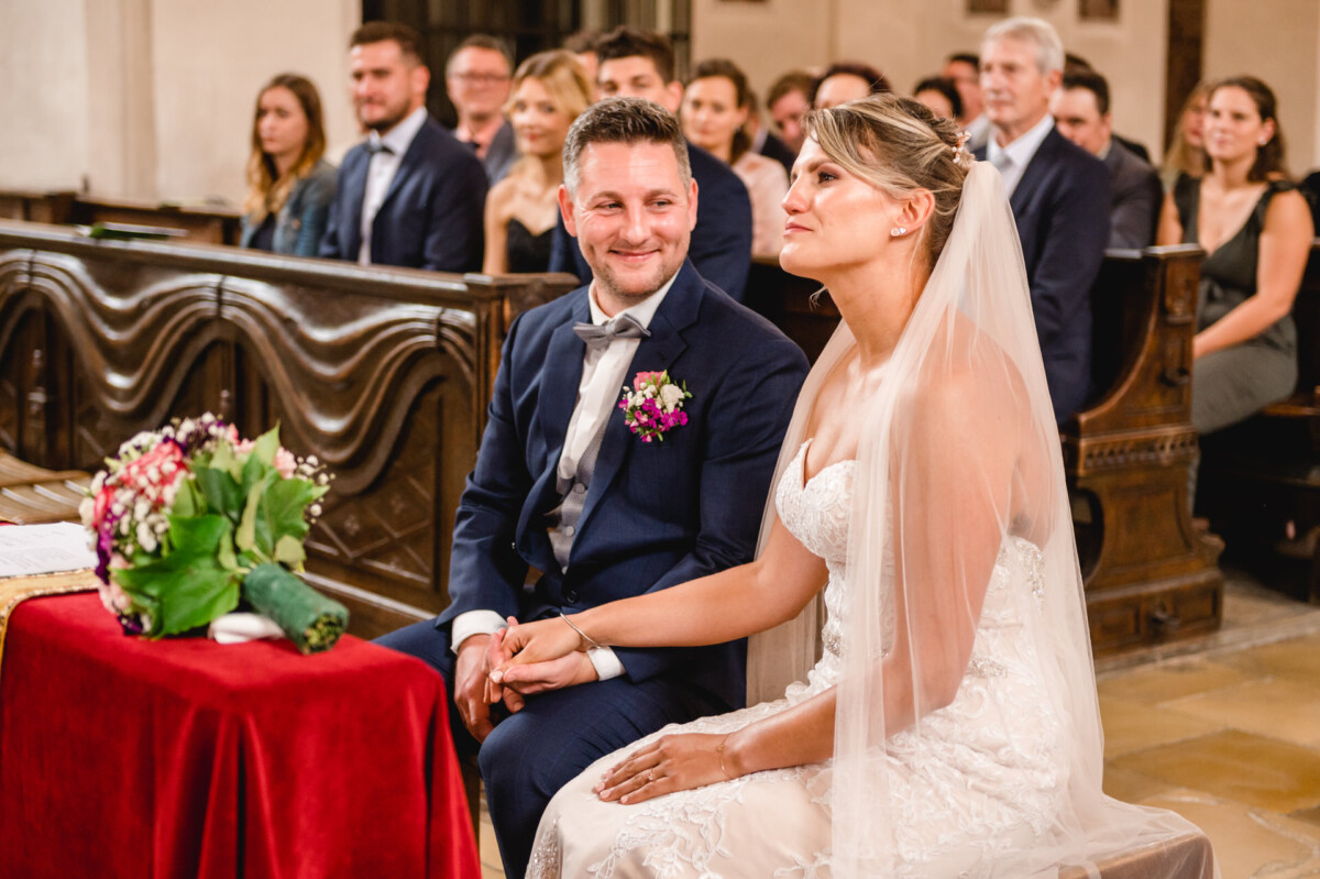 christian-lorenz.net Braut und Bräutigam sitzen Händchen haltend in einer Kirche. Die Braut trägt ein weißes Kleid und einen Schleier; der Bräutigam einen dunklen Anzug mit Fliege. Im Hintergrund sitzen Menschen.