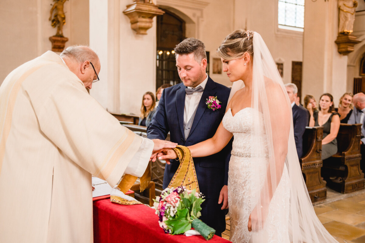 christian-lorenz.net Ein Paar nimmt an einer Hochzeitszeremonie in einer Kirche teil. Ein Geistlicher wickelt ihnen ein Tuch um die Hände. Die Braut hält einen Blumenstrauß in der Hand. Im Hintergrund sitzen Gäste.