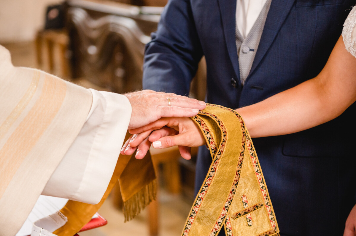 christian-lorenz.net Nahaufnahme der Hände eines Priesters, der während einer Hochzeitszeremonie ein goldenes Tuch über die Hände eines Paares legt.