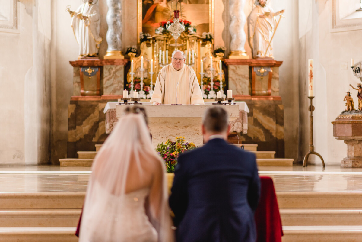 christian-lorenz.net Ein Paar kniet vor einem Altar, während ein Priester in einer mit Blumen und Kerzen geschmückten Kirche die Hochzeitszeremonie durchführt.