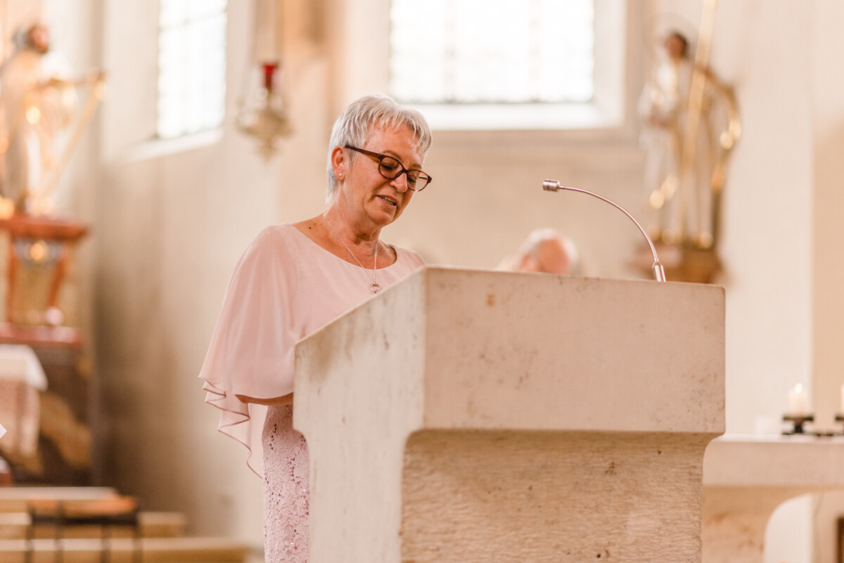 christian-lorenz.net Eine ältere Frau mit Brille spricht an einem Podium in einer Kirche, im Hintergrund sind Statuen und Fenster zu sehen.
